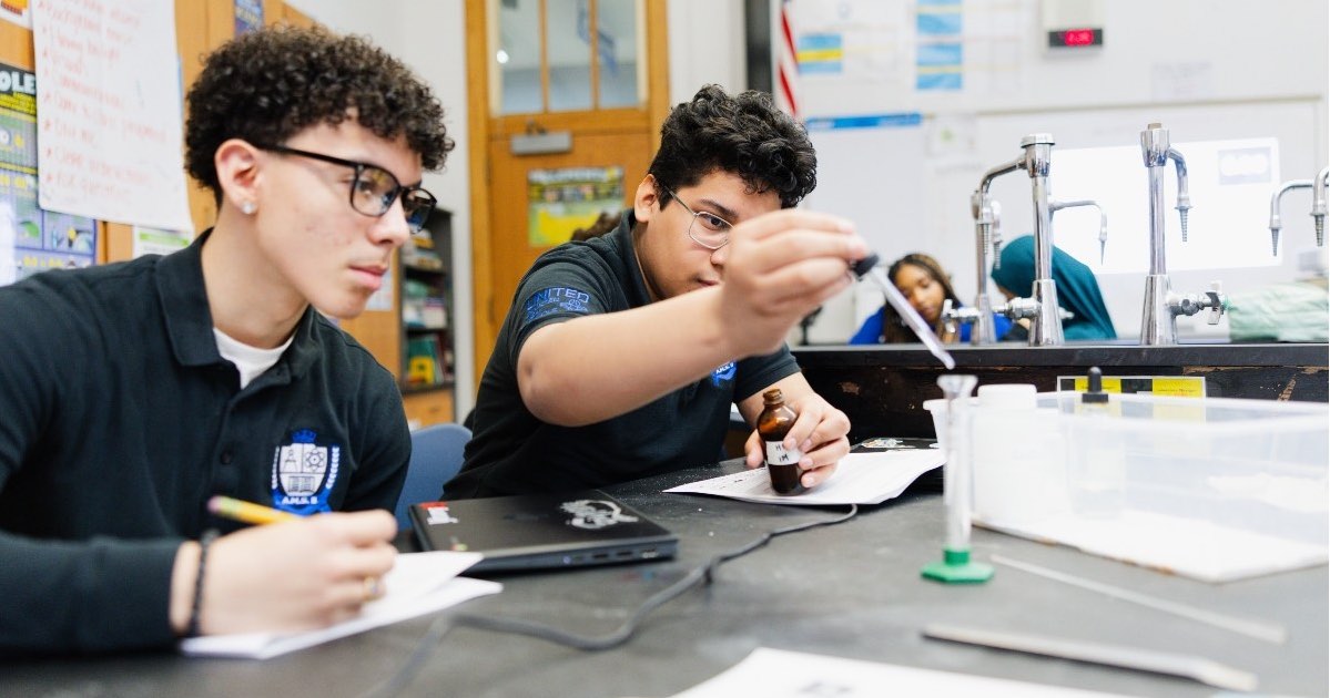 Two United Charter High School AMS II students at a lab table conduct a science experiment, with one using a dropper while the other takes notes.