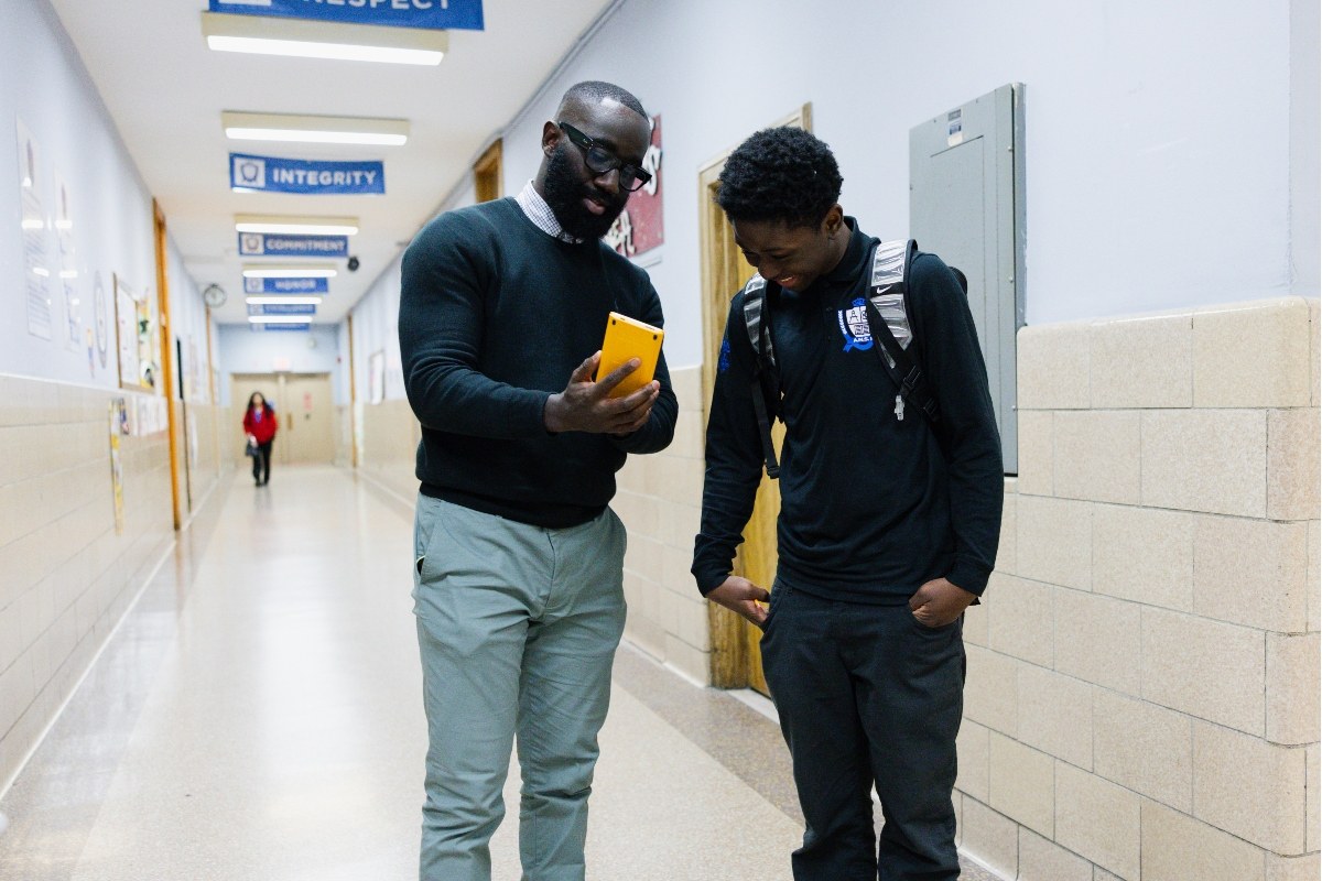 An adult and a student stand in United Charter High school 's hallway, looking at a yellow object together. The student is wearing a backpack. Blue signs hang from the ceiling in the background.