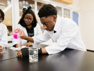 Three United Charter High School students wearing white lab coats perform a science experiment, with one student pouring a pink liquid into a graduated cylinder.